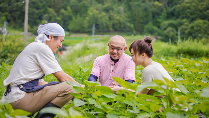 緑豊かな畑で3人の男女が集まり、作物を見ながら談笑している様子